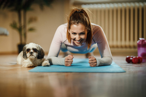 An attractive young woman doing plank exercises at home during the day supporting by her pet dog. - Fotoğraf, Görsel