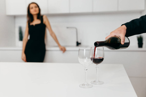 Ένα αντρικό χέρι ρίχνει κόκκινο κρασί σε ποτήρια. Γυναίκα στο παρασκήνιο - Φωτογραφία, εικόνα