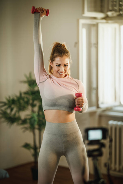 An attractive young woman doing exercises with weight at her home and using camera to record her vlog about healthy living. - Foto, Imagem
