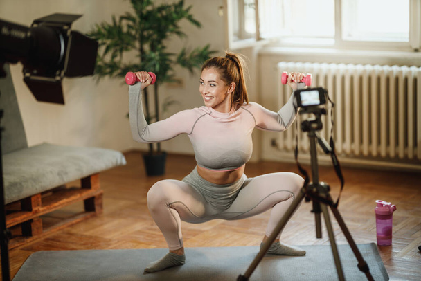 An attractive young woman doing exercises with weight at her home and using camera to record her vlog about healthy living. - Photo, Image