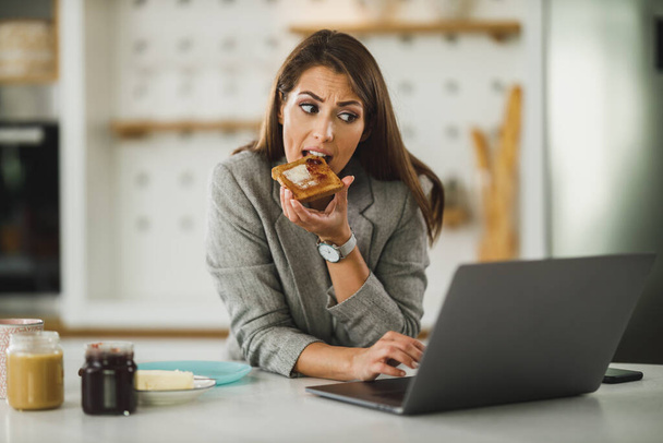 Wielozadaniowa młoda kobieta biznesu jedząca śniadanie i korzystająca z laptopa podczas przygotowywania się do pracy. - Zdjęcie, obraz