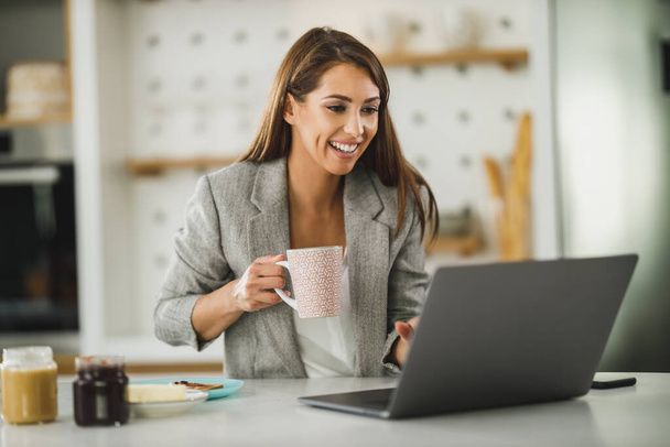 Sonriente joven mujer de negocios utilizando el ordenador portátil y beber café de la mañana en casa. - Foto, Imagen