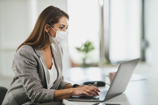 Młoda kobieta biznesu z maską ochronną siedzi sama w swoim biurze i pracuje na laptopie podczas pandemii wirusa korony. - Zdjęcie, obraz