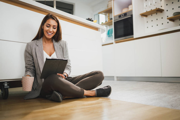 Mujer sonriente pasar el tiempo libre y el uso de la tableta digital mientras se sienta en el suelo en la cocina en su casa. - Foto, imagen