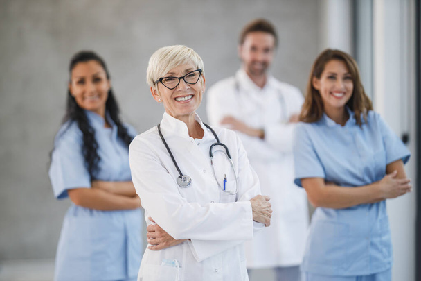 Retrato de una mujer sonriente médico senior de pie con los brazos cruzados en un pasillo del hospital con sus colegas en el fondo. - Foto, Imagen