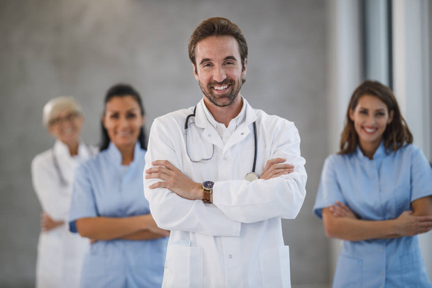 Πορτρέτο ενός χαμογελαστού γιατρού με αυτοπεποίθηση στέκεται με τα χέρια σταυρωμένα σε ένα διάδρομο του νοσοκομείου με τους συναδέλφους του στο παρασκήνιο. - Φωτογραφία, εικόνα