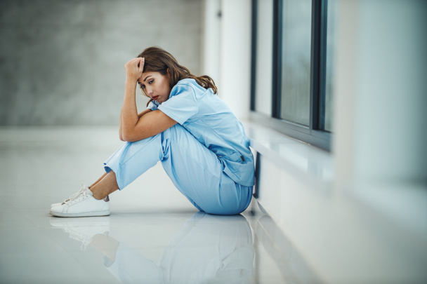 Een gestresste jonge verpleegster zit op de vloer bij een raam tijdens een snelle pauze in een lege ziekenhuisgang tijdens de Covid-19 pandemie. - Foto, afbeelding