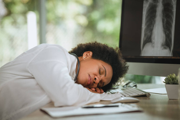 Prise de vue d'une femme africaine fatiguée allongée sur ses bras croisés au bureau alors qu'elle regardait les rayons X des poumons sur ordinateur pendant la pandémie de COVID-19. - Photo, image