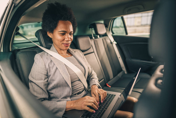 Prise de vue d'une femme d'affaires noire à succès utilisant un ordinateur portable alors qu'elle était assise sur le siège arrière d'une voiture pendant son trajet matinal. - Photo, image
