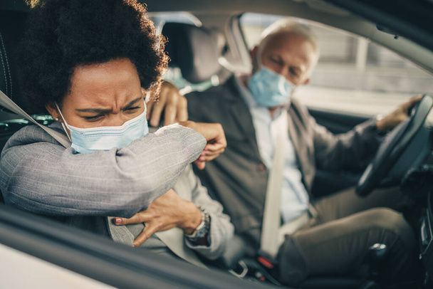 コロナウイルスのパンデミックの間彼女のシニア同僚と車で旅行中に肘で保護マスク咳を持つ成功した黒人ビジネス女性. - 写真・画像