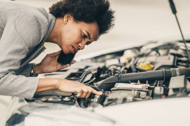 Worried black woman looking under the hood of her car while calling for help after vehicle breakdown on a roadtrip. - Photo, Image