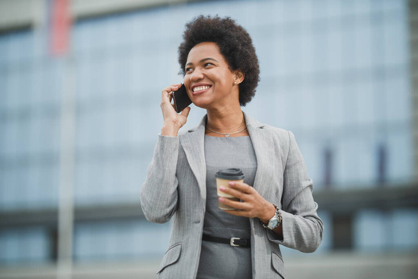Πορτρέτο μιας επιτυχημένης μαύρης επιχειρηματία που χρησιμοποιεί την εφαρμογή σε ένα έξυπνο τηλέφωνο ενώ έχει ένα διάλειμμα για καφέ μπροστά από ένα εταιρικό κτίριο. - Φωτογραφία, εικόνα