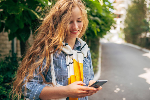 Horizontal image of the pretty female in blue shirt and pullover on the shoulders messaging on a mobile phone. A beautiful woman sending emails online on a smartphone outside. - Foto, imagen