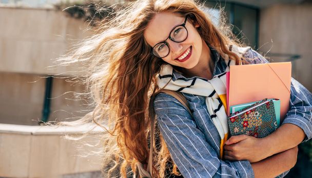 Horizontal portrait of a cheerful young student woman with red hair wearing transparent eyeglasses standing next to the college campus and carrying lots of books and folders on a sunny day. - Photo, image