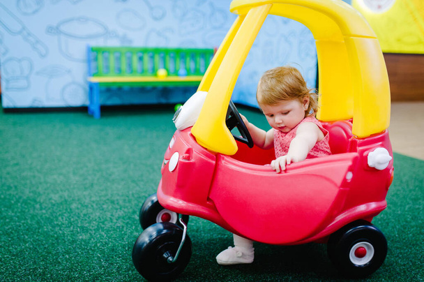 Happy children, little girl child rides on a red big car on the road. Close up. The baby is driving the car and played in a children's playroom on a birthday party. Time together at entertainment centre. - Photo, image