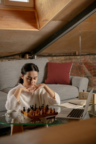 Νεαρό όμορφο κορίτσι κάθεται στο σπίτι και παίζει σκάκι σε απευθείας σύνδεση χρησιμοποιώντας φορητό υπολογιστή. - Φωτογραφία, εικόνα