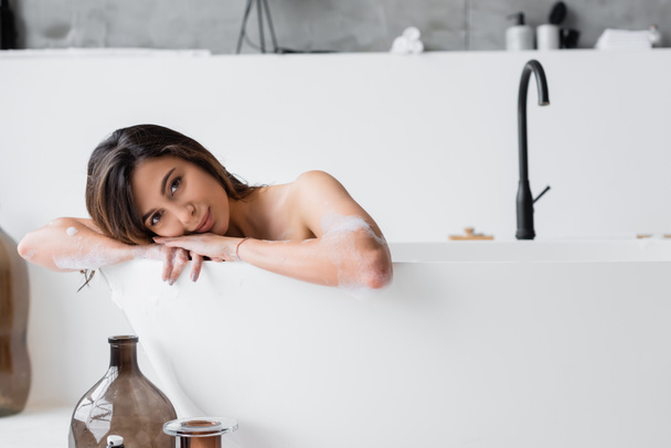 Νεαρή γυναίκα με αφρό κοιτάζει την κάμερα ενώ κάνει μπάνιο στο σπίτι  - Φωτογραφία, εικόνα