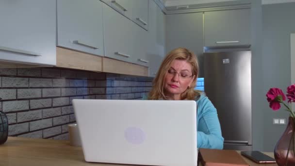 Mujer caucásica con gafas y cabello rubio se concentra mientras trabaja en la computadora en la cocina - Imágenes, Vídeo