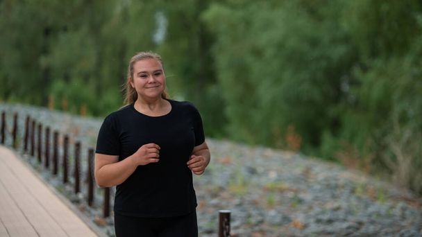 Een zwaarlijvige jonge vrouw die buiten jogt. Fat mooi lachend meisje in een zwart trainingspak is bezig met fitness voor gewichtsverlies aan het water. Een vrouw rent op een zomerdag. - Foto, afbeelding