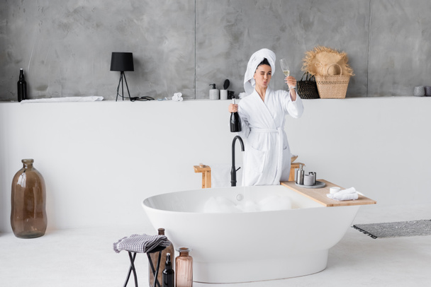 Young woman in bathrobe holding bottle and glass of champagne in bathroom  - Photo, Image