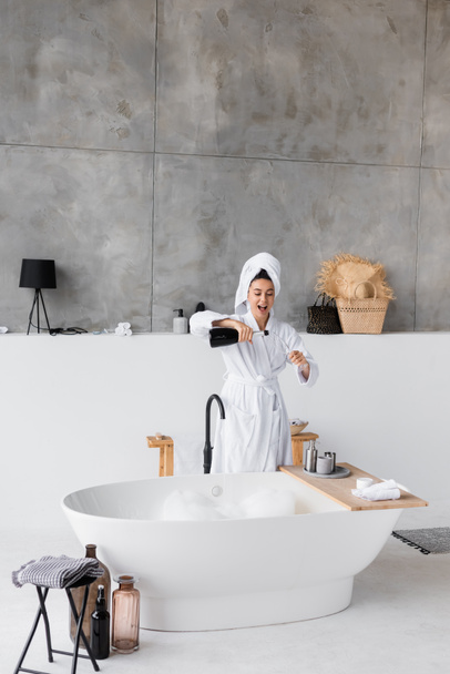 Cheerful woman pouring champagne in glass near bathtub at home  - Foto, Imagem