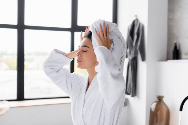 Woman in bathrobe holding white towel on head in bathroom  - Фото, изображение