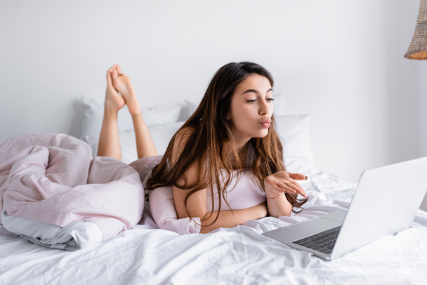 Woman blowing air kiss at laptop on blurred foreground on bed  - Foto, afbeelding