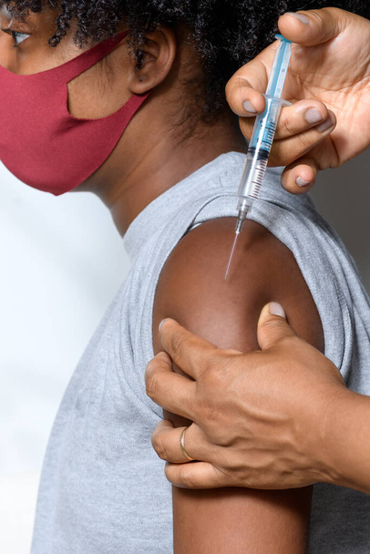 Musta teini yllään suojaava naamio vastaan covid-19 hymyillen hänen kasvonsa näyttää rokotteen tuotemerkin, eristetty valkoisella taustalla - Valokuva, kuva