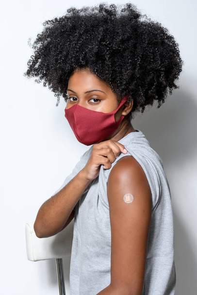black teenager wearing protective mask against covid-19 with a smile on his face shows the vaccine brand, isolated on white background - Photo, Image