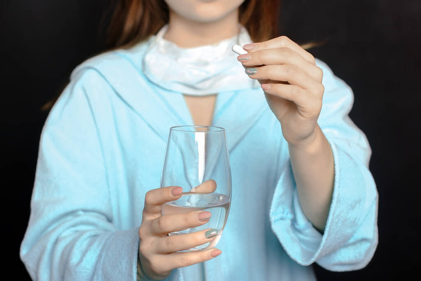 mujer joven sobre un fondo negro sosteniendo una pastilla y un vaso de agua en sus manos - Foto, imagen