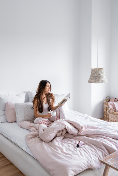 Smiling woman with coffee cup holding book on bed  - Foto, imagen