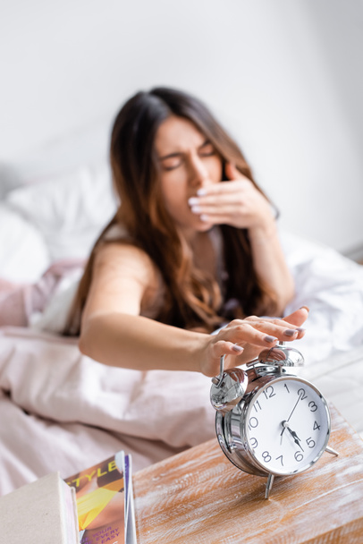 Sleepy woman on blurred background turning off alarm clock on bedside table  - Photo, Image