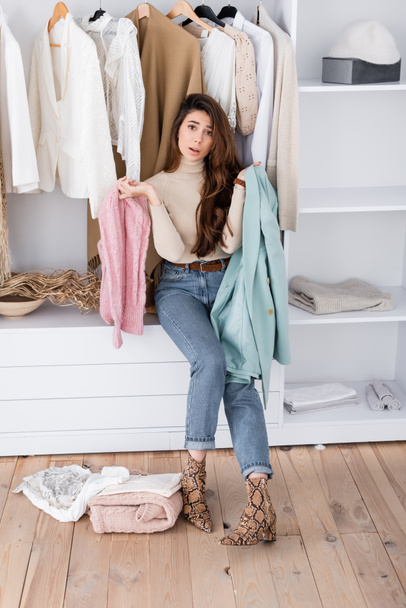 Mujer mirando a la cámara mientras sostiene la ropa cerca de estantes en el armario  - Foto, Imagen
