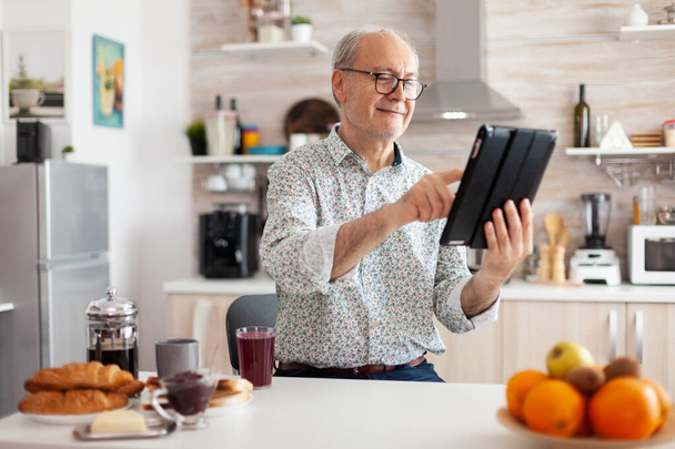 Щасливий старший чоловік шукає планшет на кухні під час сніданку
 - Фото, зображення