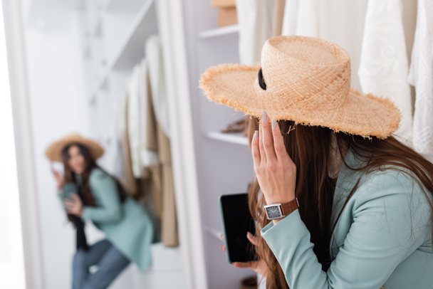 Jovem mulher de chapéu de sol segurando smartphone no fundo borrado no guarda-roupa  - Foto, Imagem