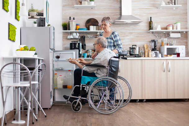 Zdravotně postižený starší muž na invalidním vozíku brát vejce z chladničky pomáhá své ženě - Fotografie, Obrázek