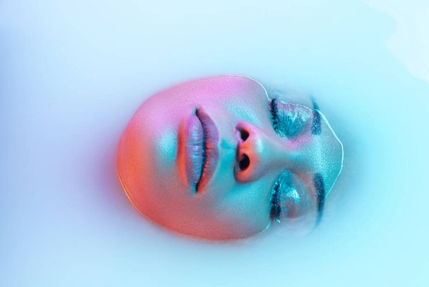 Beautiful female face in the milk bath with soft glowing in blue-pink neon light - Photo, image