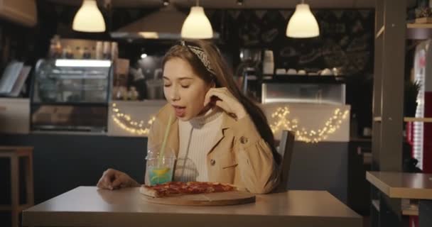 Uma bela jovem loira senta-se em um café em uma mesa com fatias de pizza em uma tábua de madeira e bebe um coquetel azul com limão através de uma palha - Filmagem, Vídeo
