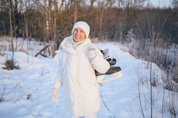 Ευτυχισμένη ηλικιωμένη ώριμη γυναίκα σε λευκό ζεστό outwear παίζει με παγοπέδιλα σε ηλιόλουστο χιονισμένο χειμώνα σε εξωτερικούς χώρους. Συνταξιούχοι υγιείς άνθρωποι διακοπές χειμερινές δραστηριότητες, ενεργός τρόπος ζωής έννοια - Φωτογραφία, εικόνα
