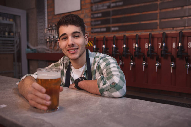 Young bartender smiling joyfully, serving glass ov delicious light beer, copy space - Фото, изображение