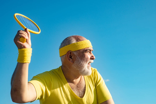 Elderly man practicing tennis on blue sky background. Senior man tennis player serving. - Фото, зображення