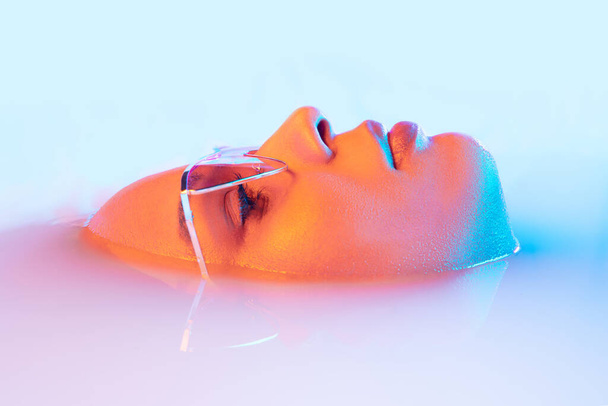 Beautiful female face in the milk bath with soft glowing in blue-pink neon light - Фото, изображение