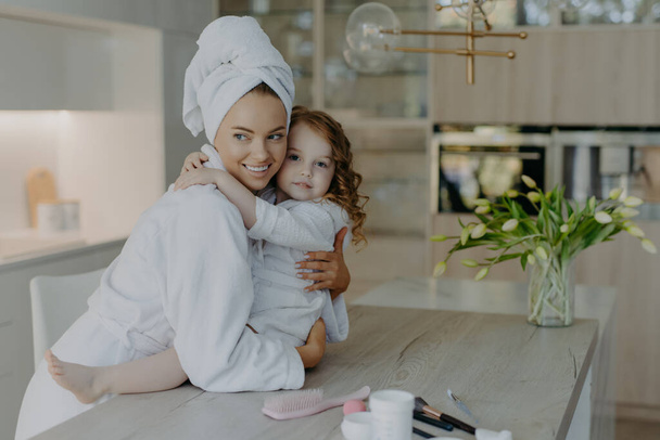 Photo of young mother and daughter embrace and express love to each other pose in modern apartment wear soft white bath robes going to have cosmetic procedures have healthy skin. Time for beauty - Foto, Imagen