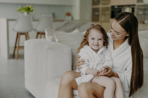 Beautiful brunette affectionate mother holds small curly haired cute dauhter in dressing gown pose in cozy room against modern apartment interior. Mom and little girl at home after taking shower - Foto, imagen