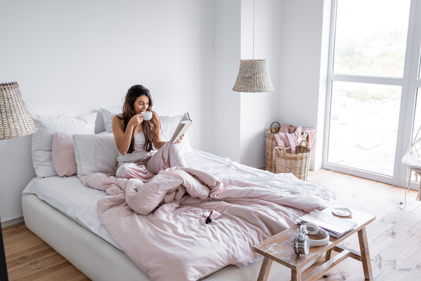 Mujer bebiendo café y leyendo libro en dormitorio moderno  - Foto, imagen
