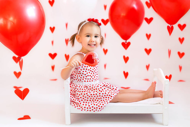 Dziecko trzyma czerwone kulki na Walentynki. Dziewczyna je słodycze w kształcie serca. Dziecko w białej sukience z sercem. - Zdjęcie, obraz