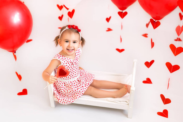Baby hält rote Kugeln zum Valentinstag in der Hand. Ein Mädchen isst ein Bonbon in Herzform. Ein Kind im weißen Kleid mit Herzen. - Foto, Bild