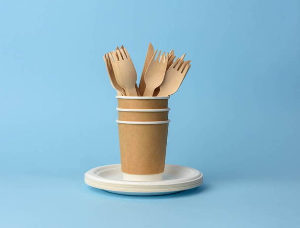 papír pohár, fehér tányérok, fa villák és kések kék alapon. Műanyag elutasítási koncepció, nulla hulladék  - Fotó, kép