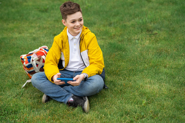 Školák sedí na trávníku s chytrým telefonem v ruce. Moderní teenager ve žluté bundě - Fotografie, Obrázek