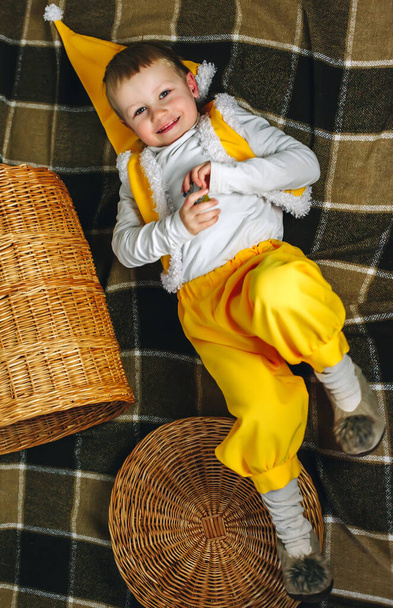 Little boy 4 years old lay on home plaid in yellow gnome elf outfit with cap and straw basket in short pants, turtleneck shirt, tights and fluffy slippers, warm cozy colors - Photo, Image